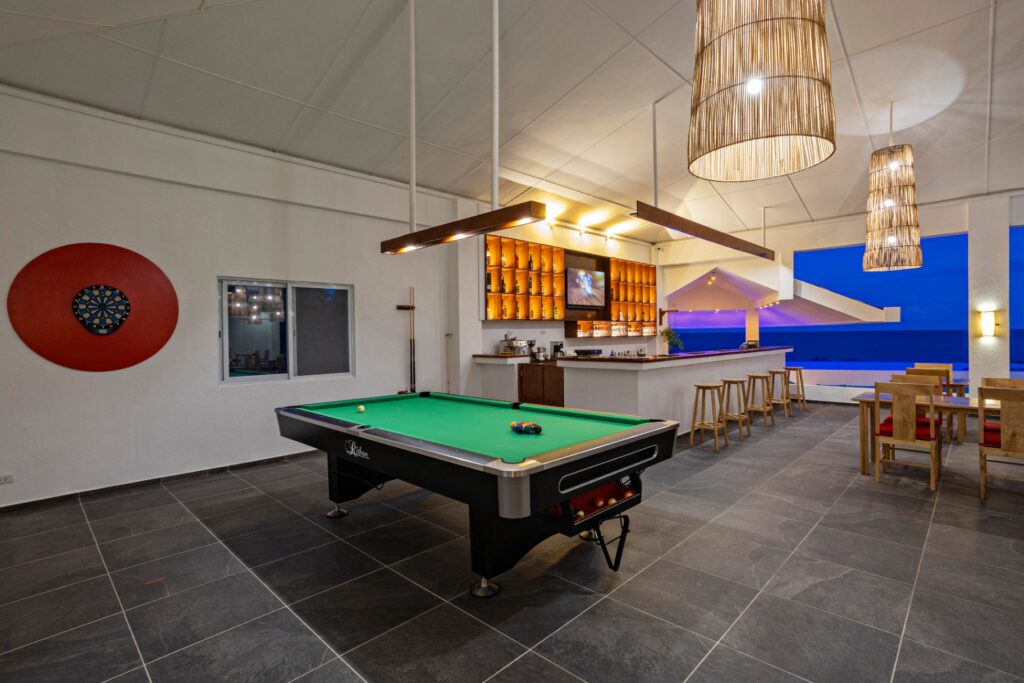 A modern game room with a pool table, dartboard, bar area, and dining tables overlooks a blue-lit outdoor view—an inviting retreat for international visitors exploring Boljoon Cebu or relaxing after a day at Dive Spot Asia.